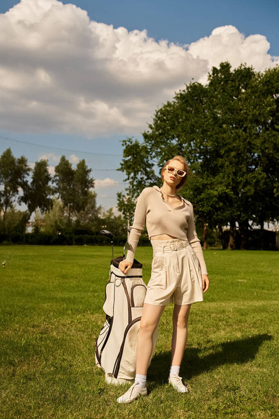Μια εκλεπτυσμένη νεαρή γυναίκα στέκεται με χάρη σε ένα πεδίο με μια τσάντα του γκολφ, απολαμβάνοντας την ύπαιθρο με έναν αέρα εκλεπτυσμένης κομψότητας. - Φωτογραφία, εικόνα