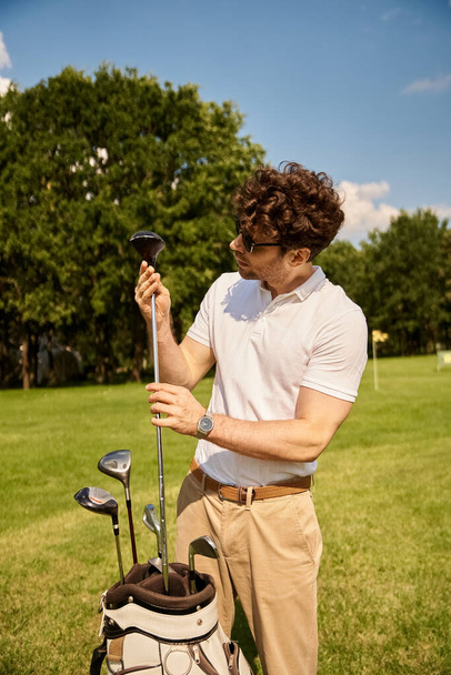 Tyylikäs mies tyylikkäissä vaatteissa pitää golfkassia rehevällä vihreällä kentällä arvostetulla golfmailalla, johon sisältyy yläluokan vapaa-aika.. - Valokuva, kuva