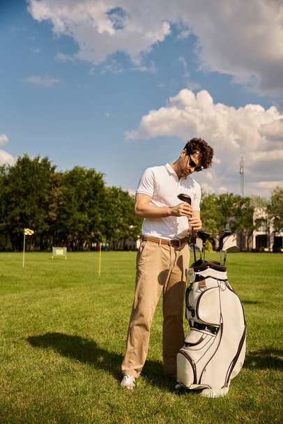 エレガントな服装の男は,高級ゴルフクラブで緑豊かな畑のゴルフバッグの横に優雅に立っています. - 写真・画像