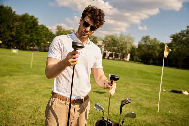 Homem elegante em roupas elegantes fica ao ar livre, segurando com confiança um taco de golfe em suas mãos em um campo verde exuberante. - Foto, Imagem
