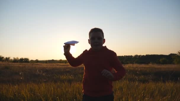 Unbekümmert joggt ein kleiner Junge mit einem Papierflugzeug durch die Wiese. Nettes kleines Kind, das über die Wiese rennt und ein Spielzeugflugzeug über dem Hintergrund des Sonnenuntergangs startet. Konzept eines Kindheitstraums. Nahaufnahme. - Filmmaterial, Video