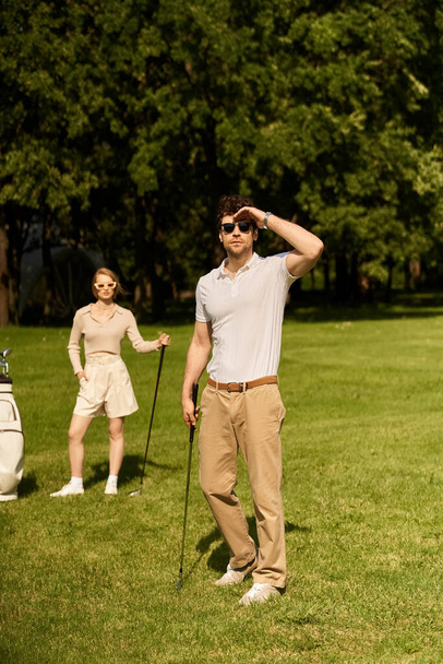 Ένα νεαρό ζευγάρι με κομψή ενδυμασία παίζει γκολφ σε ένα πράσινο γήπεδο στο πάρκο, απολαμβάνοντας μια χαλαρή μέρα μαζί. - Φωτογραφία, εικόνα