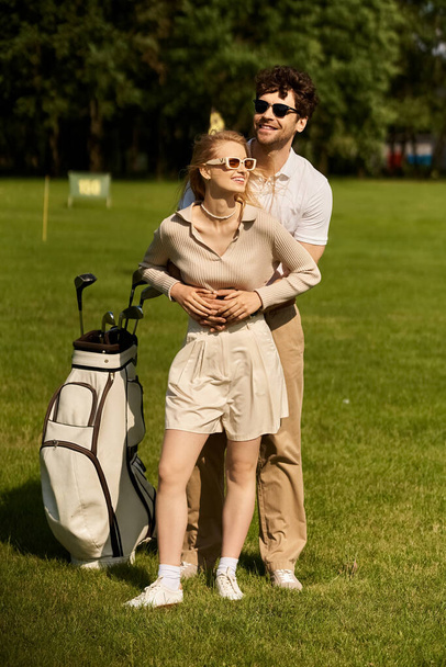 Ένα νεαρό ζευγάρι με κομψή ενδυμασία στέκεται μαζί σε ένα καταπράσινο γήπεδο γκολφ, αγκαλιάζοντας την αριστοκρατική ατμόσφαιρα. - Φωτογραφία, εικόνα