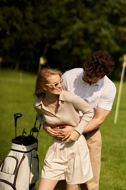 Ένα νεαρό ζευγάρι με κομψή ενδυμασία μοιράζονται μια ζεστή αγκαλιά σε ένα παρθένο γήπεδο γκολφ, που περιβάλλεται από την ομορφιά του πράσινου τοπίου. - Φωτογραφία, εικόνα