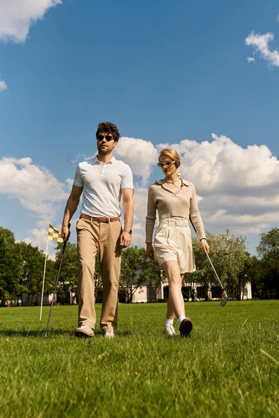 Молодой человек и женщина в элегантной одежде прогуливаются вместе на пышном зеленом поле для гольфа, наслаждаясь высококлассной активностью на открытом воздухе. - Фото, изображение