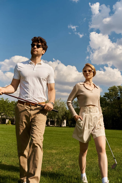 Un homme et une femme élégants se promènent à travers un champ animé, tenant des clubs de golf sous le ciel clair d'un après-midi tranquille. - Photo, image