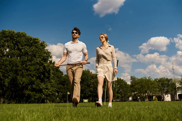 エレガントな服装のスタイリッシュな若いカップルは,ゴルフクラブの設定で緑豊かな畑を散策します. - 写真・画像