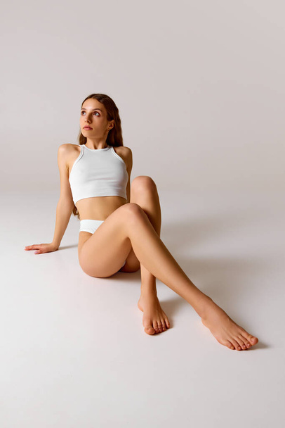Young, barefoot woman sitting on floor with crossed legs with well-kept, smooth skin against white studio background. Concept of beauty and health, depilation, anti-cellulite program, dieting. - Photo, Image