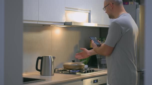 Mężczyzna w ubraniu casual smażenie kurczaka na patelni w kuchni i nagrywać wideo na blogu na smartfonie komórkowym. Hobby, przygotowanie domu żywności, nowoczesne wnętrze, koncepcja gotowania - Materiał filmowy, wideo