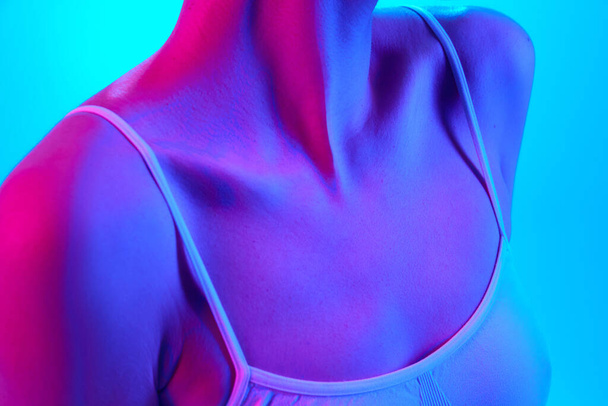 Close-up of well-kept, smooth female body, neckline, breast in sports bra in neon light against gradient background. Concept of beauty and health, cosmetology products for sun protection, recreation. - Photo, Image