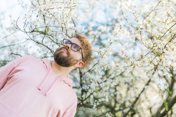 Knappe man buiten portret op achtergrond kersenbloesems of appelbloesems en blauwe lentehemel. Millennial generatie man en nieuwe mannelijkheid - Foto, afbeelding