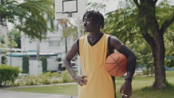 Portret wesołego czarnego gracza streetballa trzymającego piłkę podczas pozowania do kamery na placu zabaw - Materiał filmowy, wideo