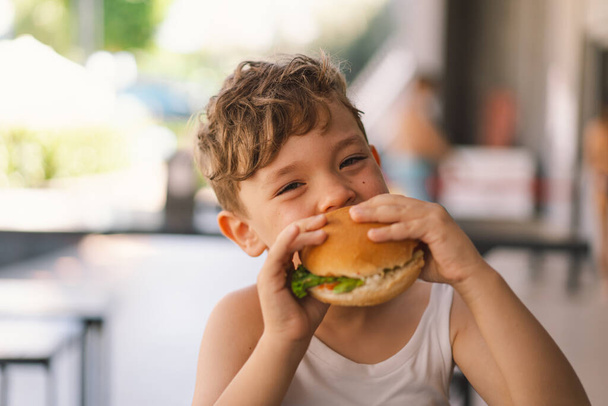 テーブルでサンドイッチとフレンチフライを食べる小さな少年. 彼は一方でサンドイッチとフレンチフライに焦点を当てているように見えます. 男の子は屋外でファーストフードを食べる. - 写真・画像