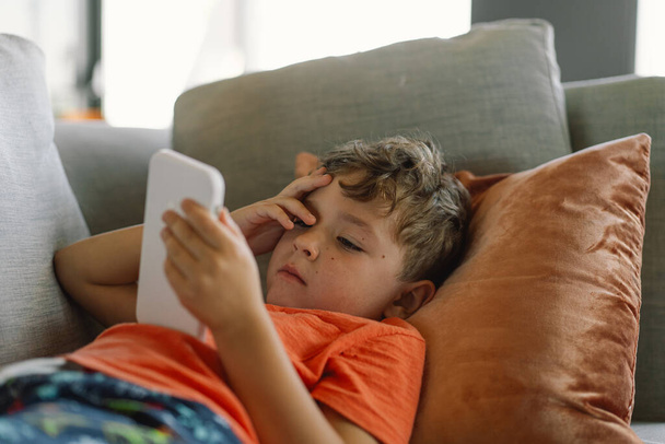 Мальчик с вьющимися волосами удобно лежит на сером диване с подушками, глубоко погруженный в игру по телефону. неторопливый день в помещении. - Фото, изображение