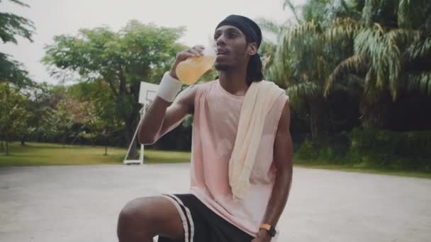 Retrato medio largo del deportista afroamericano tomando refrescante bebida de limón y sonriendo a la cámara en la cancha al aire libre - Imágenes, Vídeo