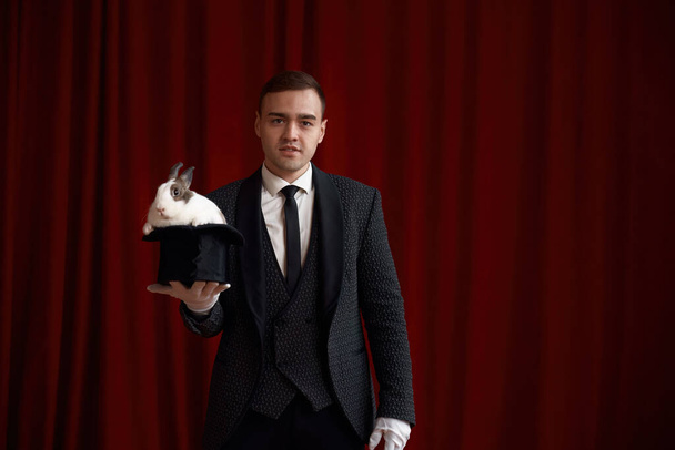 Young handsome magician holding top hat with cute fluffy rabbit entertaining performance illusion trickery on theater stage - Photo, image