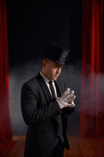 Magicien homme élégant portant costume élégant debout en fumée sur scène en regardant le public. Jeune artiste showman contre rideau draperie rouge vue de côté - Photo, image