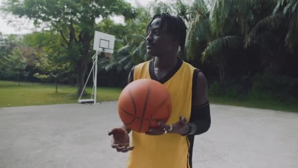 Ritratto medio di giocatore di basket nero in abbigliamento sportivo giallo in attesa del suo amico sul campo e sorridente alla macchina fotografica - Filmati, video
