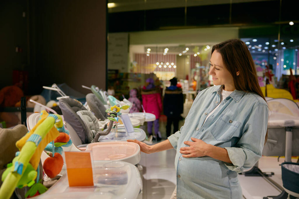Zwangere vrouw kiest nieuwe stoel voor babyvoeding uit het assortiment in de winkel. Goederen voor pasgeboren kinderen in het winkelcentrum - Foto, afbeelding
