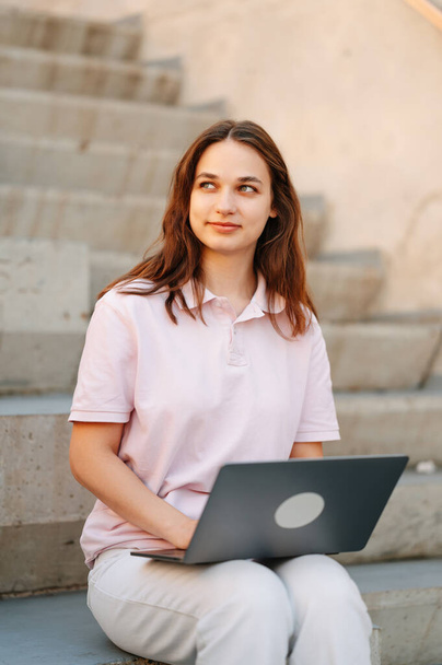 Jovem alegre sorridente estudante do sexo feminino está sentado ao ar livre escadas trabalhando no laptop. - Foto, Imagem
