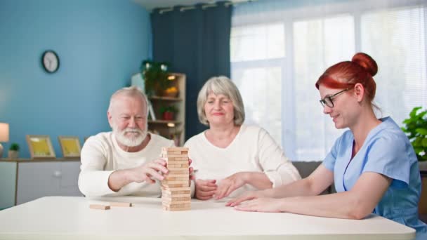 γυναίκα κοινωνικός λειτουργός παίζει επιτραπέζιο παιχνίδι με γέρος και ηλικιωμένη γυναίκα κτίριο πύργο από ξύλινα μπλοκ, ενώ κάθεται στο τραπέζι στο σπίτι, τα χέρια κοντά - Πλάνα, βίντεο
