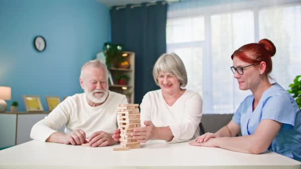 Забота о престарелых, молодая работница весело играет в настольные игры с пожилым мужчиной и женщиной, сидящих за столом в комнате - Кадры, видео