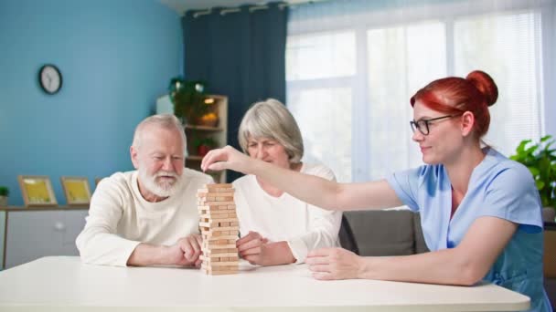 настільна гра, літня жінка і чоловік разом з молодим соціальним працівником в медичній формі весело будують вежу з дерев'яних блоків на столі, сидячи в затишній кімнаті - Кадри, відео