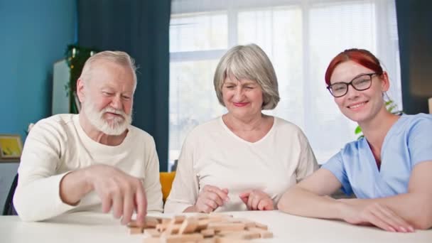 portret pracowniczki socjalnej w mundurze medycznym wraz ze starszą parą małżeńską bawiącą się grając w gry planszowe siedząc przy stole w pokoju, uśmiechając się i patrząc w kamerę - Materiał filmowy, wideo