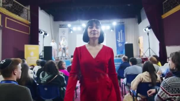 Жінка в червоній сукні стоїть у конференц-залі, аудиторії та сцені на задньому плані. Концепція публічного виступу та участі у заходах з копіювальним простором для дизайну та друку. Член залученої аудиторії - Кадри, відео