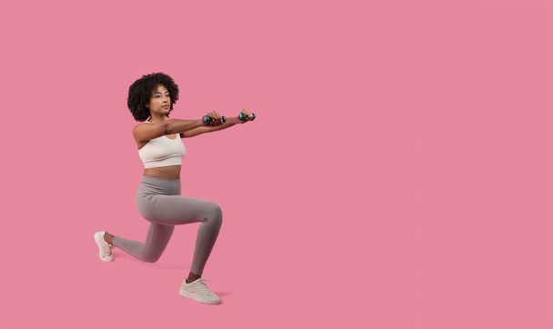 A dynamic sporty African American lady captured mid lunge holding dumbbells, showing strength and agility, isolated on pink - Photo, Image