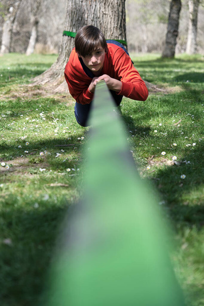 Niño concentrado mirando a la cámara mientras está acostado en slackline atado al tronco y arrastrándose con la ayuda de las manos en el bosque verde a la luz del día - Foto, imagen