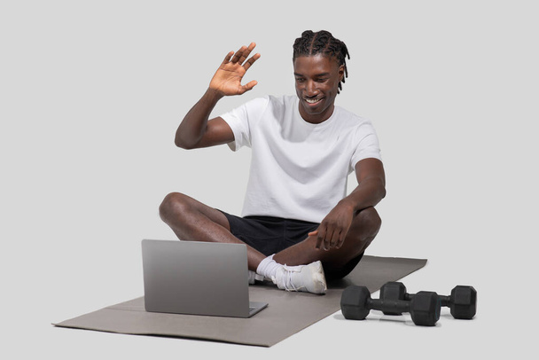 Um homem atlético preto está envolvido em uma chamada de vídeo em seu laptop, enquanto em um tapete de ginástica, com halteres nas proximidades, capturado isolado no branco - Foto, Imagem