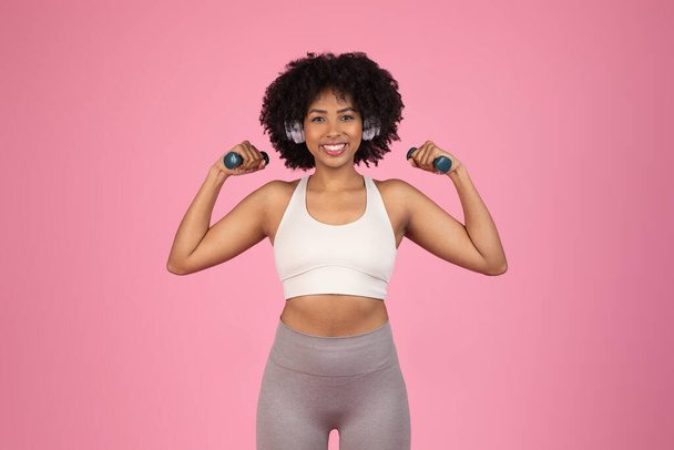 Fröhliche Afroamerikanerin lässt die Muskeln spielen, die Hanteln in der Hand, hört Musik, repräsentiert Kraft und Freude an Fitness, isoliert auf rosa - Foto, Bild