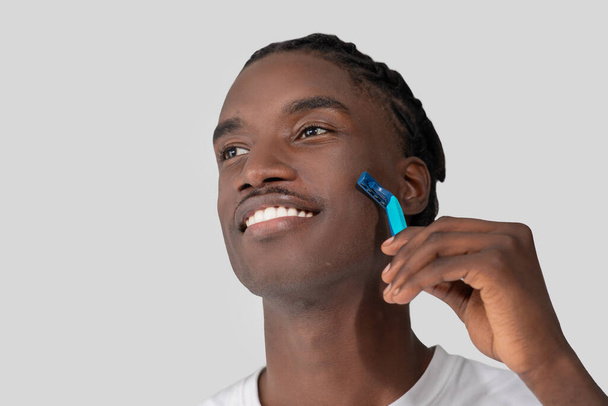 Een close-up van een vrolijke jonge Afro-Amerikaanse man met gevlochten haar, die zijn gezicht scheert met een blauw scheermes tegen een witte achtergrond - Foto, afbeelding