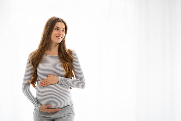 Una donna incinta contenuto in una camicia a righe e pantaloni grigi tiene la pancia e guarda a lato con un sorriso gentile, spazio copia - Foto, immagini