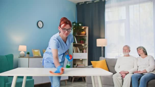 jonge vrouwelijke werknemer van een verpleeghuis helpt met het schoonmaken van een getrouwd stel, wast meubels met handschoenen met behulp van wasmiddelen - Video