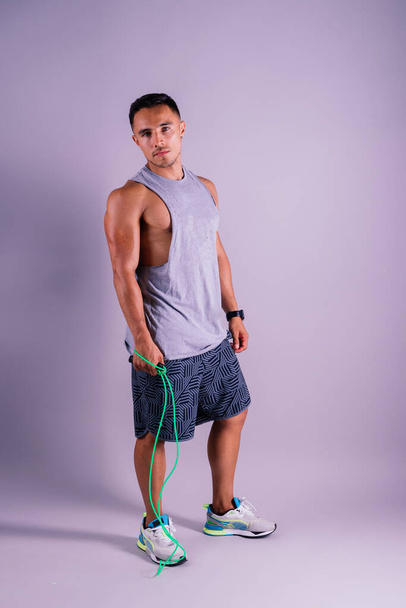 A man with a grey tank top is holding a green jump rope with his elbow bent and waist twisted, showing off his toned chest and wrist strength - Photo, Image