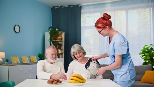 aidant un couple âgé, une jeune infirmière médicale prend soin d'un homme et d'une femme âgés verse de l'eau chaude dans des tasses tout en prenant une collation à table dans la chambre - Séquence, vidéo