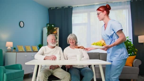 pomoc starším, ženy sociální pracovnice pečující o starší osoby v lékařské uniformě pomáhá staré ženě a muži s domácími pracemi a dává jídlo na stůl - Záběry, video
