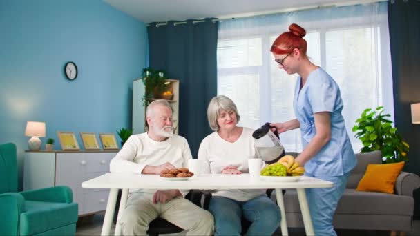zorgzame vrouwelijke maatschappelijk werker verzorgen van oude man en vrouw zetten fruit en koekjes op tafel voor hen tijdens de lunch - Video