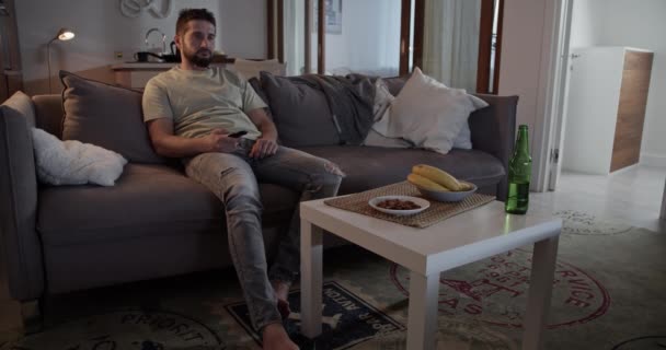 Osamělý vousatý dospělý muž v ležérním oblečení přepínání televizních kanálů pomocí dálkového ovládání při ležení na pohovce v obývacím pokoji doma - Záběry, video