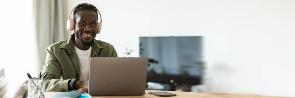 Positive black man in wireless headphones working online on laptop at home office, looking and smiling at camera, panorama with copy space. Happy guy communicating online on portable pc - Photo, Image
