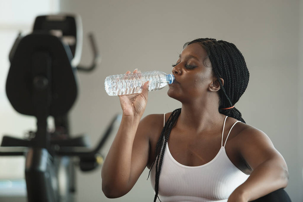 Atlético saludable fuerte hembra afroamericana más ropa deportiva de tamaño beber agua en botella después de hacer ejercicio en el gimnasio, Agua potable refresca el cuerpo. concepto de relajación y ejercicio. - Foto, imagen