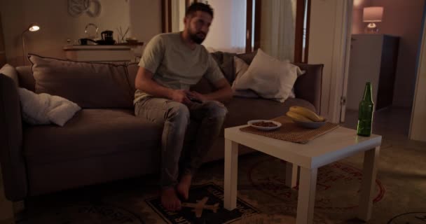 Yksinäinen parrakas aikuinen mies rento vaatteita vaihtamalla TV-kanavia kaukosäätimellä kun makaa sohvalla olohuoneessa kotona - Materiaali, video