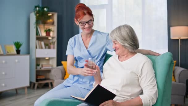 retrato de una joven enfermera trae un vaso de agua a una anciana, sonriendo y mirando a la cámara - Metraje, vídeo