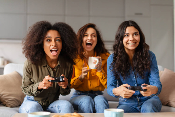 Три многорасовых девушки молодые люди наслаждаются видеоигры вместе, выражая волнение в домашней обстановке - Фото, изображение