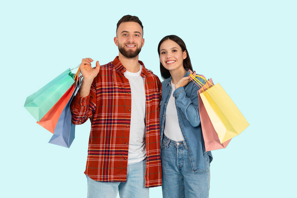 Χαμογελαστό ζευγάρι με πολλαπλές πολύχρωμες τσάντες για ψώνια, παρουσιάζοντας έναν καταναλωτικό τρόπο ζωής Zoomer σε μπλε φόντο - Φωτογραφία, εικόνα