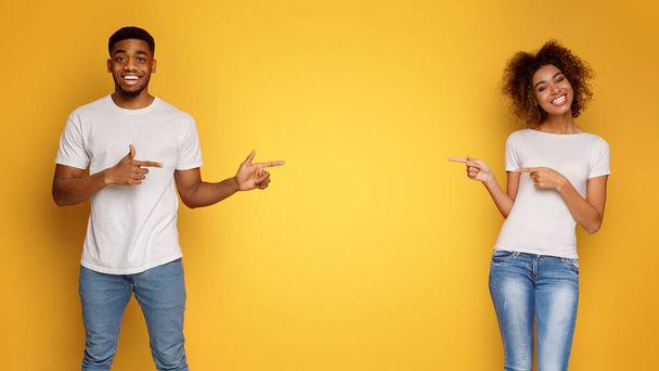 Look over here Cheerful african-american man and woman smiling and cheerfully pointing at empty space between them, orange background - Photo, Image