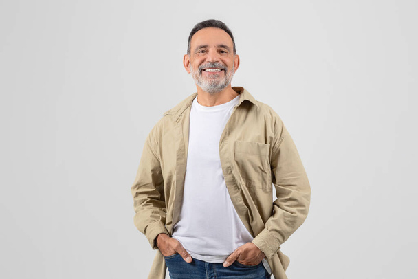 Portrait d'un vieil homme joyeux, vêtu d'une veste et d'un jean, les mains tranquillement reposées dans ses poches, isolées sur fond blanc - Photo, image