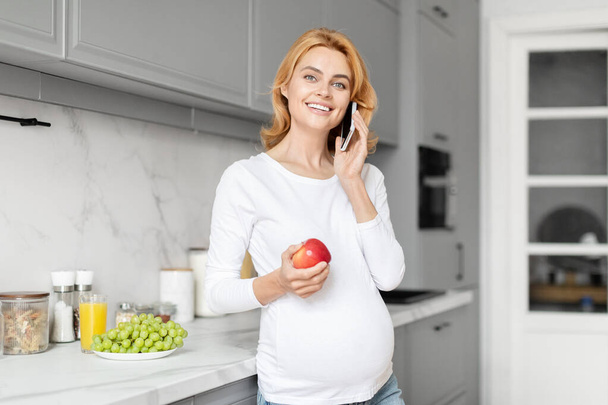 Una donna incinta europea felice parla al telefono nella sua cucina, circondata da elementi nutrizionali della cucina - Foto, immagini